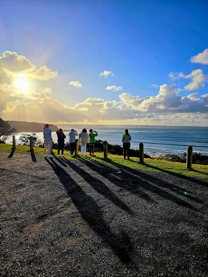 A group of people standing on a gravel path by the sea, with the ocean and a clear sky in the background. 3