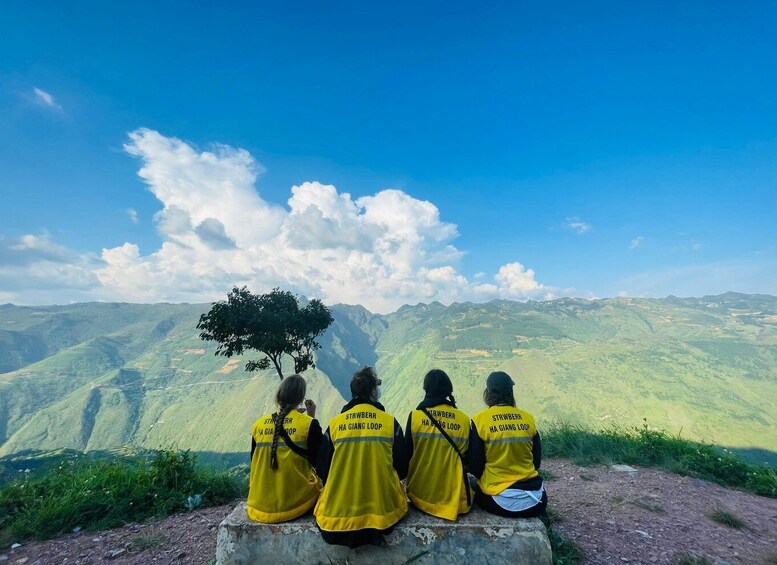 Four people in yellow vests sitting on a ledge overlooking a mountainous landscape. 1