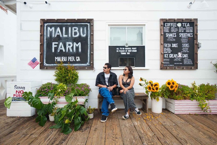 A couple sitting on a bench outside a cafe named Malibu Farm in Malibu, CA, with a menu board listing coffee, tea, shakes, smoothies, fresh juice, sodas, ice cream, and cake. 4