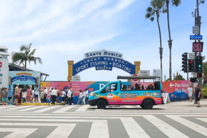 A colorful bus with 'Hollywood Bus Tours' and 'Pier Access This Way' signs, crossing a street with pedestrians and palm trees. 3