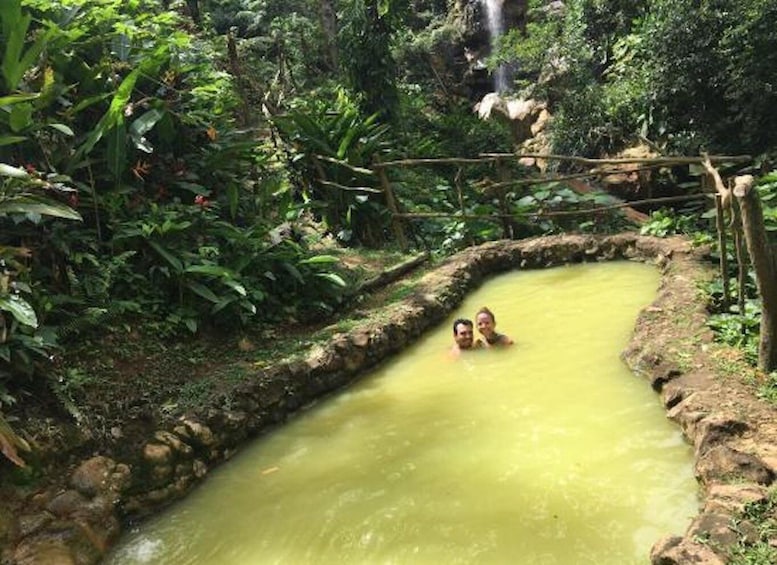 A natural hot spring with two people in the water surrounded by lush greenery. 3