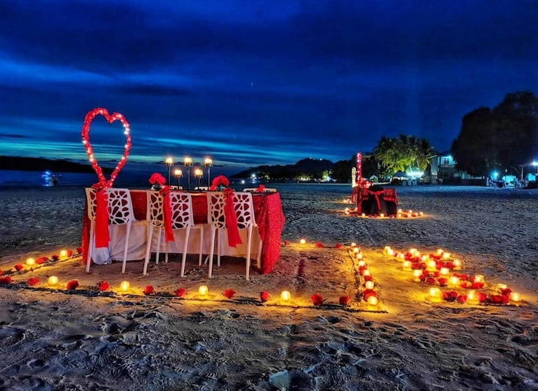 A romantic setup on a beach at night with a table decorated with red and white linens, a heart-shaped light, and candles. 3