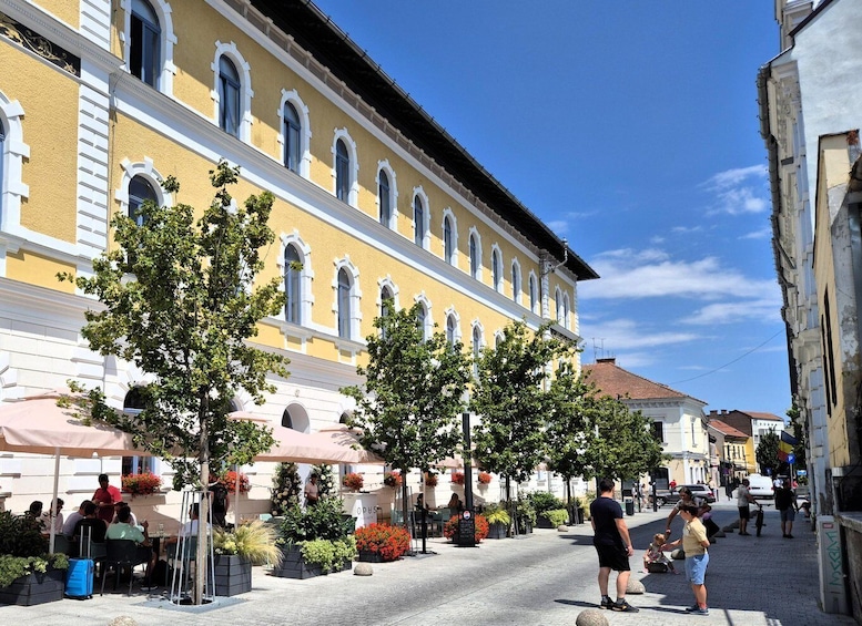 A street with a yellow building, outdoor cafes, and people walking. 4