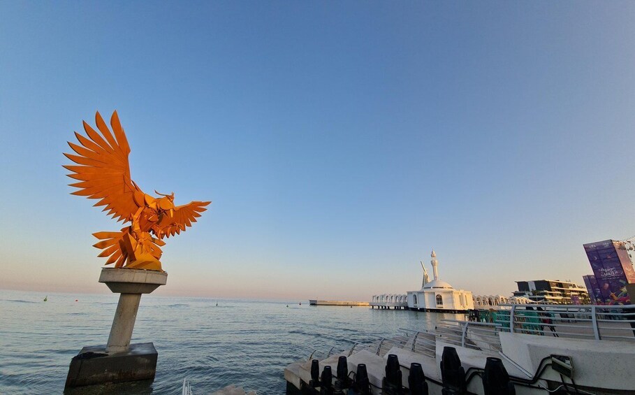 A statue of an eagle on a pedestal by the water, a boat docked nearby, and a mosque with a minaret in the background. 3