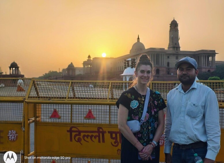 Two people standing in front of a yellow barrier with text, with a historic building and a tower in the background during sunset. 3
