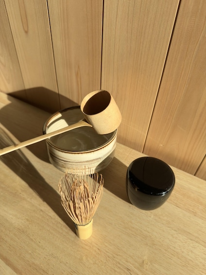 A wooden table with a tea set, including a black bowl, a white bowl with a brown rim, and a wooden whisk. 3