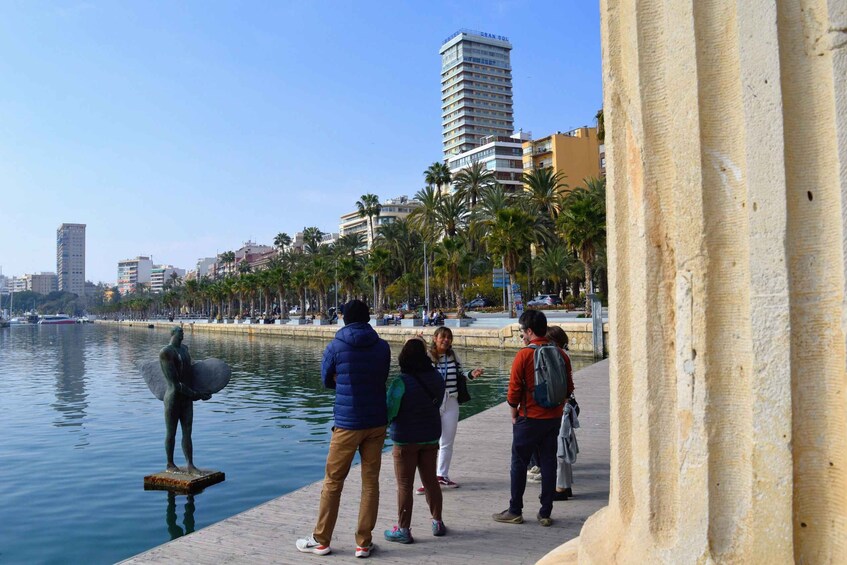 A waterfront promenade with people walking, a statue, and palm trees. 1