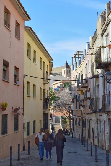 A narrow street lined with pastel-colored buildings, a clear blue sky, and a few pedestrians. 3