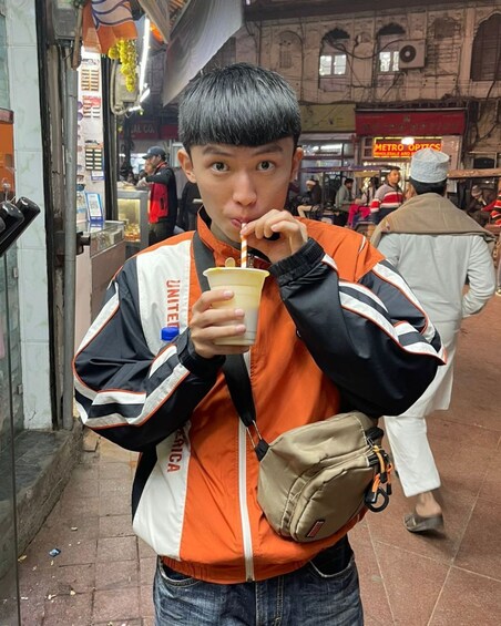 A person in an orange and black jacket holding a drink with a straw, standing on a street with shops and people in the background. 3