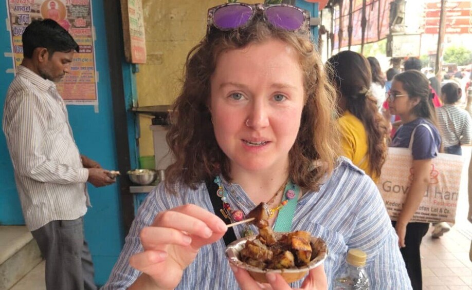 A person eating food with a plate in hand, standing in an outdoor setting with a blue wall and people in the background. 2