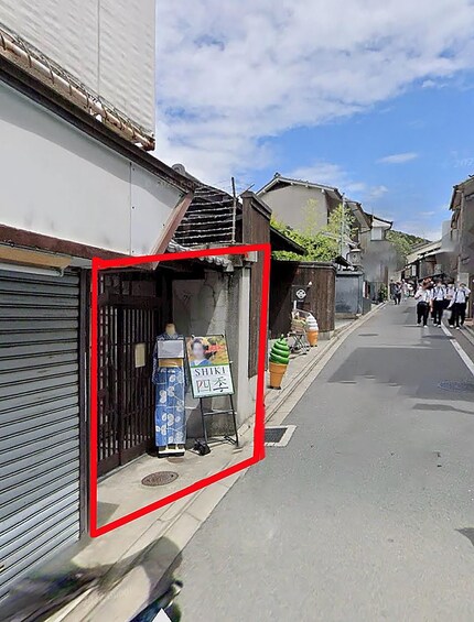 A street with traditional buildings, a closed shop with a signboard, and pedestrians in the background. 5