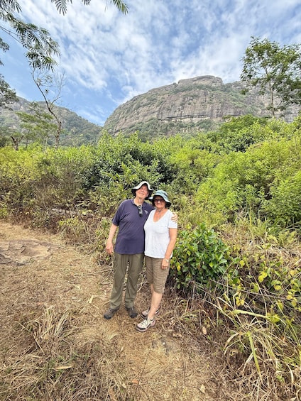 Two people standing in a natural setting with dense vegetation and a rocky cliff in the background. 3