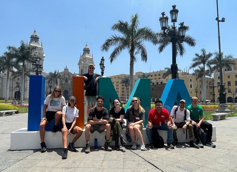 A group of people sitting on benches in front of a large 'A' sculpture in a plaza with palm trees and historic buildings in the background. 1