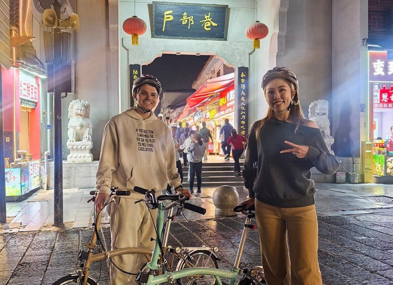 Two people standing in front of a traditional Chinese gate with a lion statue, holding bicycles. 4