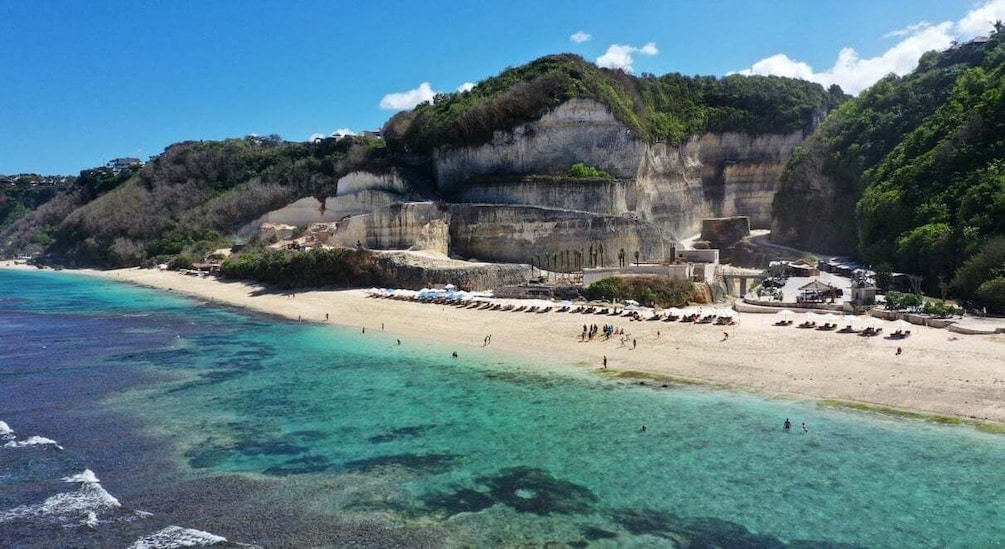 A coastal beach with clear turquoise waters, sandy shore, and a cliff with vegetation. 1