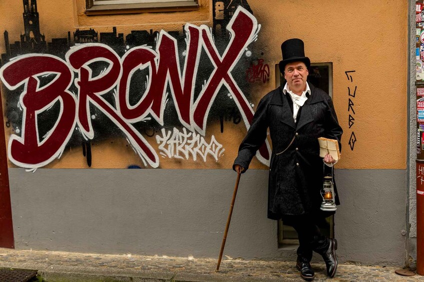 A man in a black coat and top hat holding a cane and a lantern, standing in front of a wall with graffiti that reads "BRONX". 2