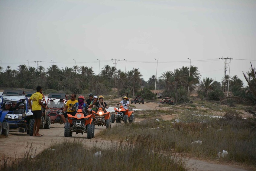 People on orange off-road vehicles, a blue vehicle, and a car parked on a dirt road with palm trees in the background. 4