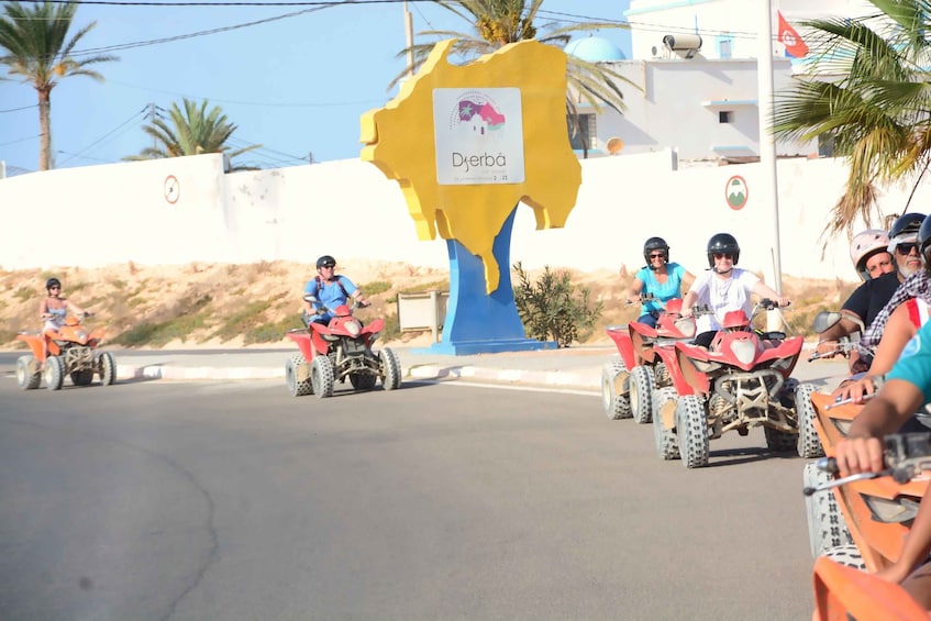 A group of people riding quad bikes on a road with a yellow and blue sign that reads "Djerba". 1