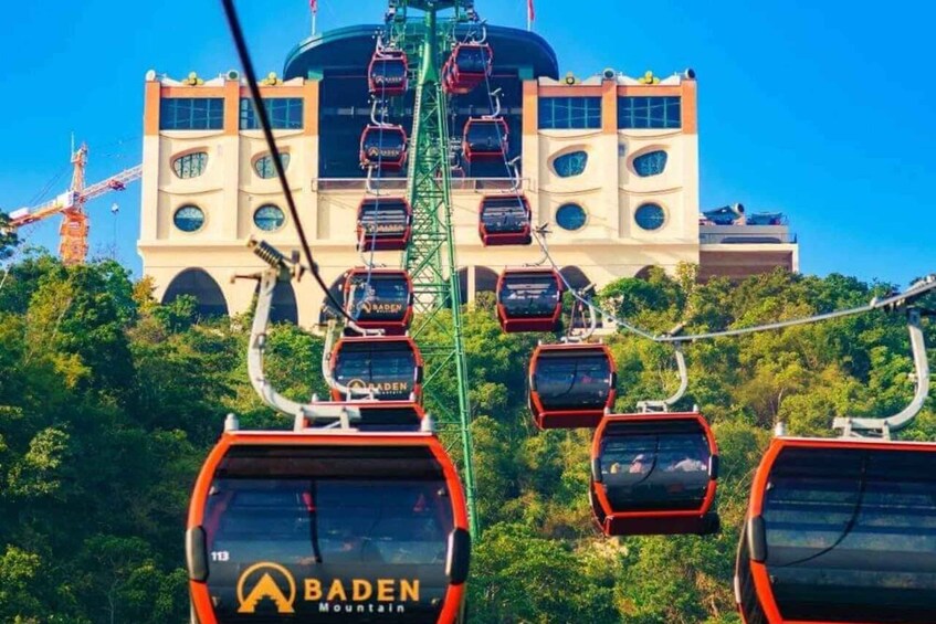 A cable car system with red cars labeled "BADEN Mountain" against a backdrop of a large building and a construction crane. 1