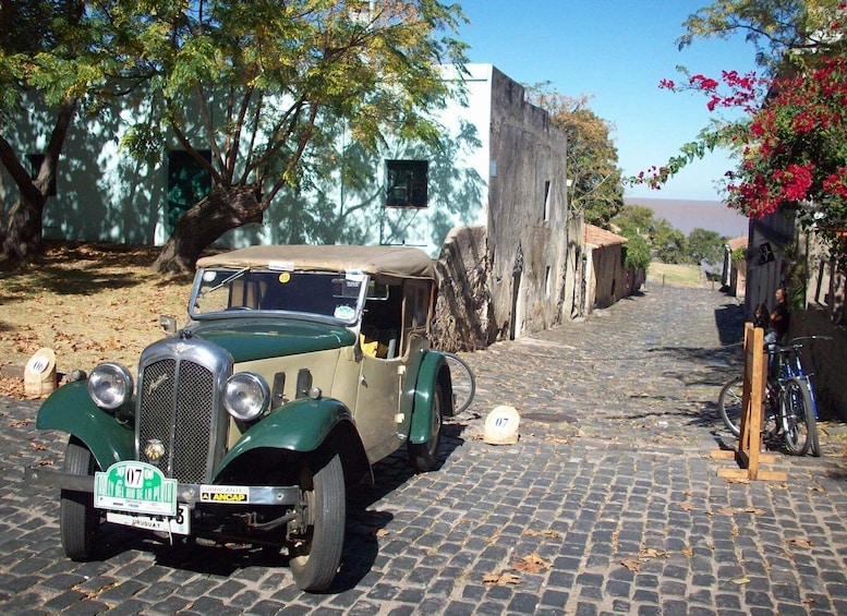 A vintage green car parked on a cobblestone street, surrounded by trees and buildings. 2