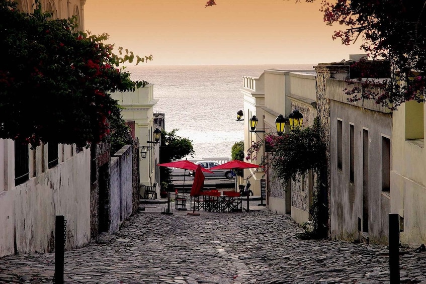 A cobblestone street leading to a body of water, with buildings on either side and red umbrellas at the end. 1