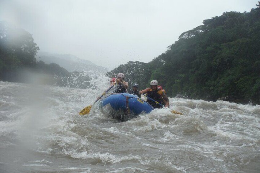 Two people navigating a blue raft through turbulent river rapids surrounded by dense forest. 4