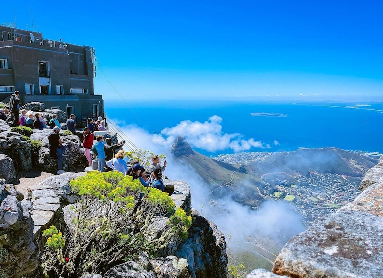 A rocky overlook with people taking photos, a building with antennas, and a view of a coastal city and ocean. 5
