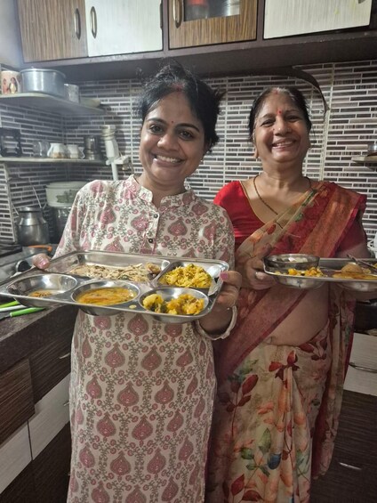 Two women holding trays of food in a kitchen with wooden cabinets and a tiled backsplash. 3
