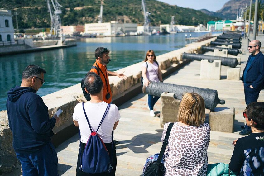 A group of people standing by a waterfront with a row of cannons and a bridge in the background. 5