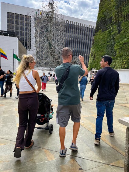 A group of people walking in an urban plaza with buildings and a green wall in the background. 2
