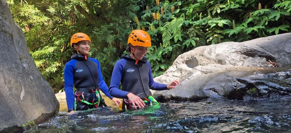 Two people in blue and black wetsuits with orange helmets standing in a river surrounded by rocks and greenery. 2