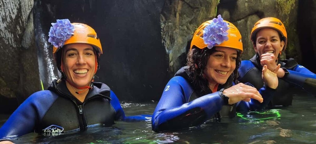 Two people in blue wetsuits and orange helmets in a water body with rocky surroundings. 1