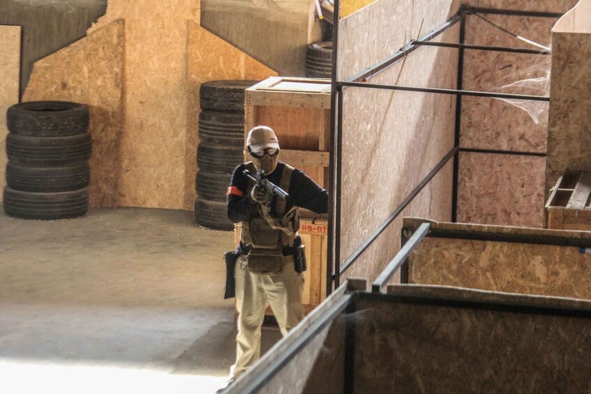A person in tactical gear holding a rifle in an indoor shooting range with wooden obstacles and tire stacks. 2
