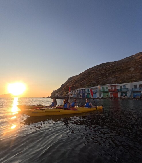 A sunset over a calm sea with a yellow kayak carrying four people. In the background, there are colorful buildings along the shore. 5