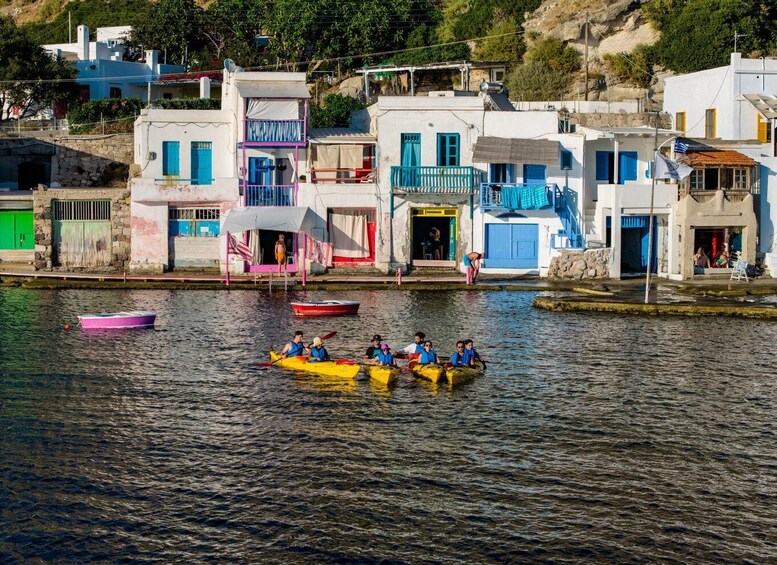 A group of people in yellow kayaks on a calm body of water, with colorful buildings and a rocky hillside in the background. 4