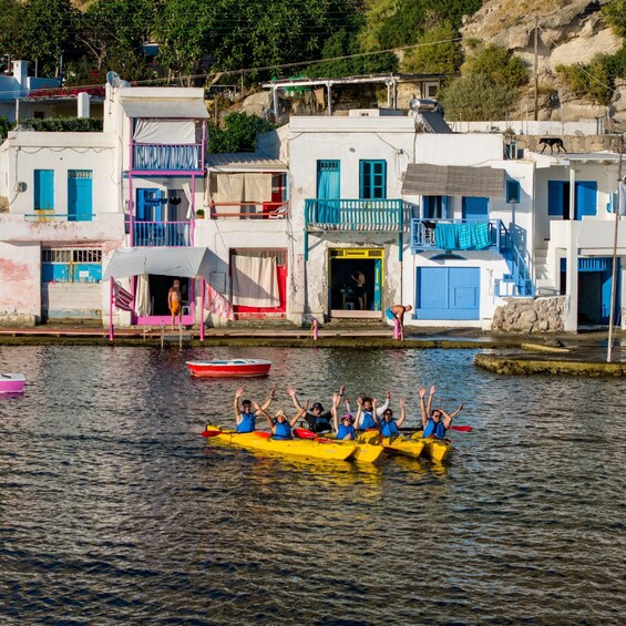 A group of people in a yellow kayak on a calm river, with colorful buildings in the background. 1