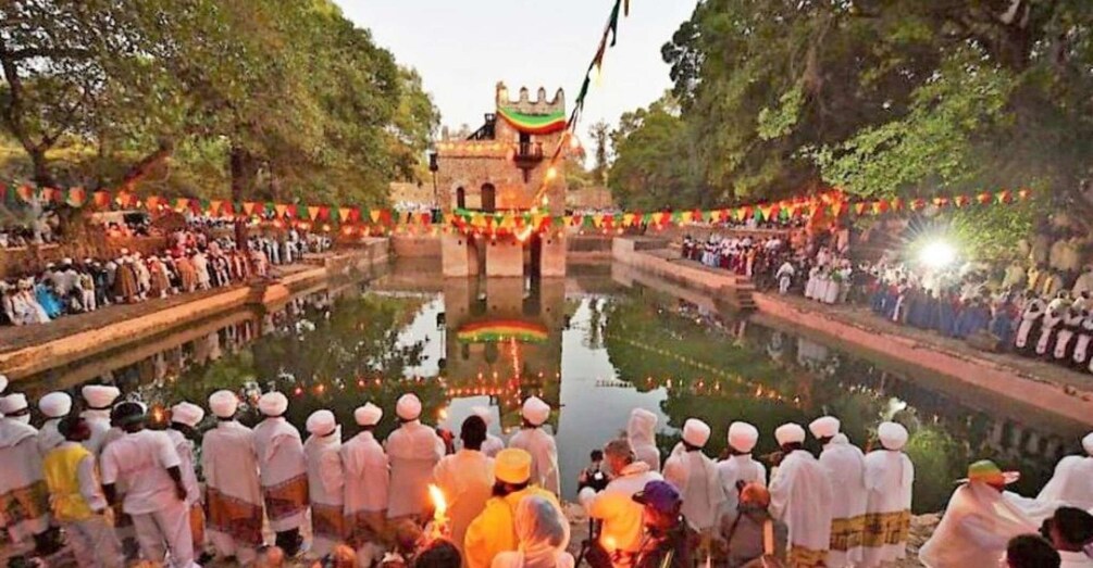 A group of people in white attire gathered around a pond, with a building in the background adorned with colorful decorations. 1