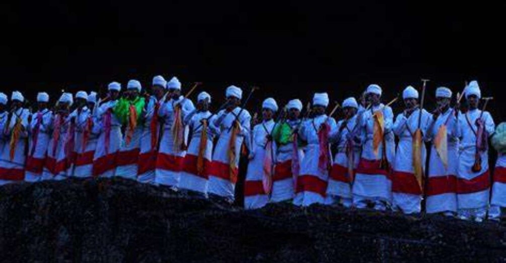 A group of people in white robes with red sashes, standing in a line outdoors at night. 5