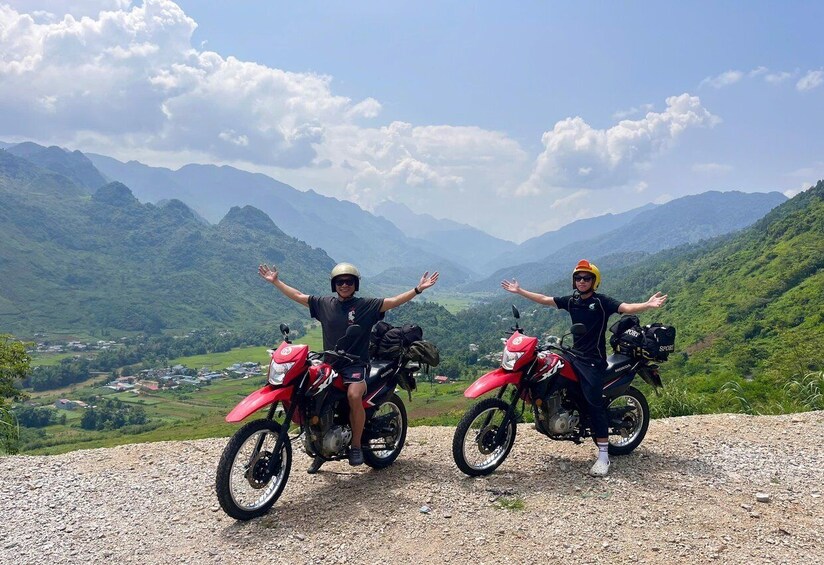 Two people on motorcycles with their arms raised, overlooking a mountainous landscape. 3