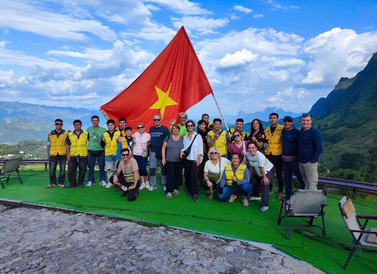 A group of people posing with a flag, standing on a green platform with a mountainous landscape in the background. 4