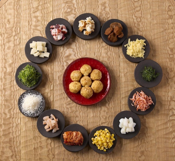 A wooden table with various food items arranged in a circular pattern around a central red plate of round pastries. 1