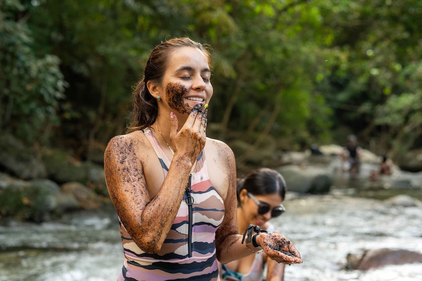A person covered in mud near a flowing river with rocks and greenery in the background. 2