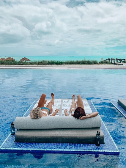 Two people relaxing on a floating lounge chair in a pool with a view of the beach and ocean. 1
