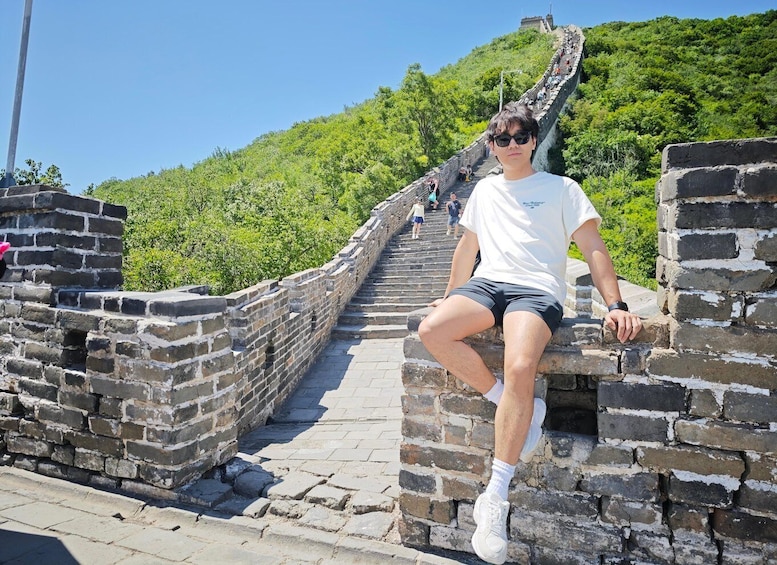 A person sitting on a stone wall of the Great Wall of China, with lush green hills in the background. 1