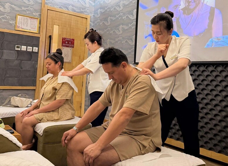 A massage therapy session with clients seated on a massage table and therapists attending to them. 2