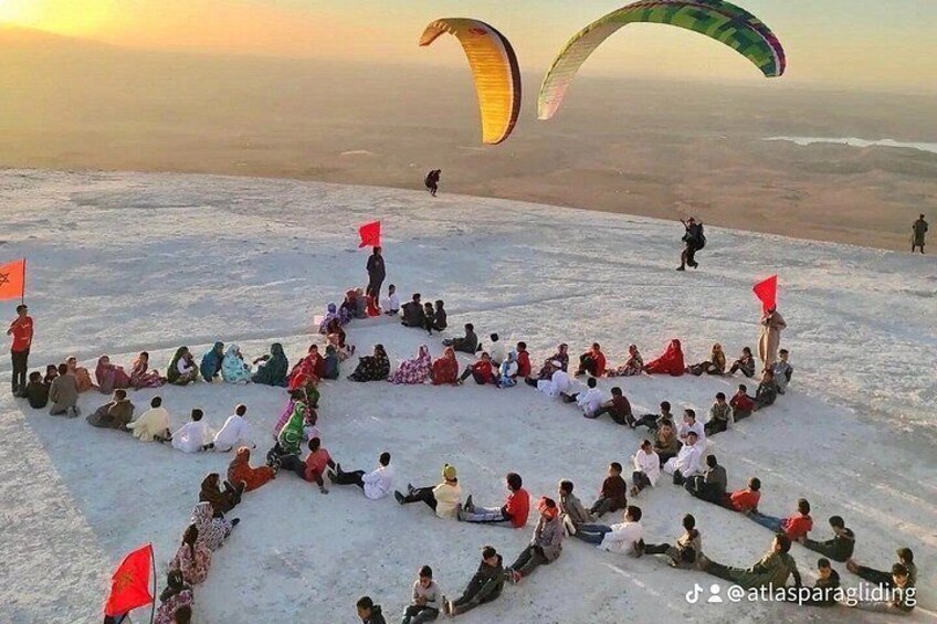 A group of people sitting in a large star formation on a snowy mountain with kites flying in the background. 2