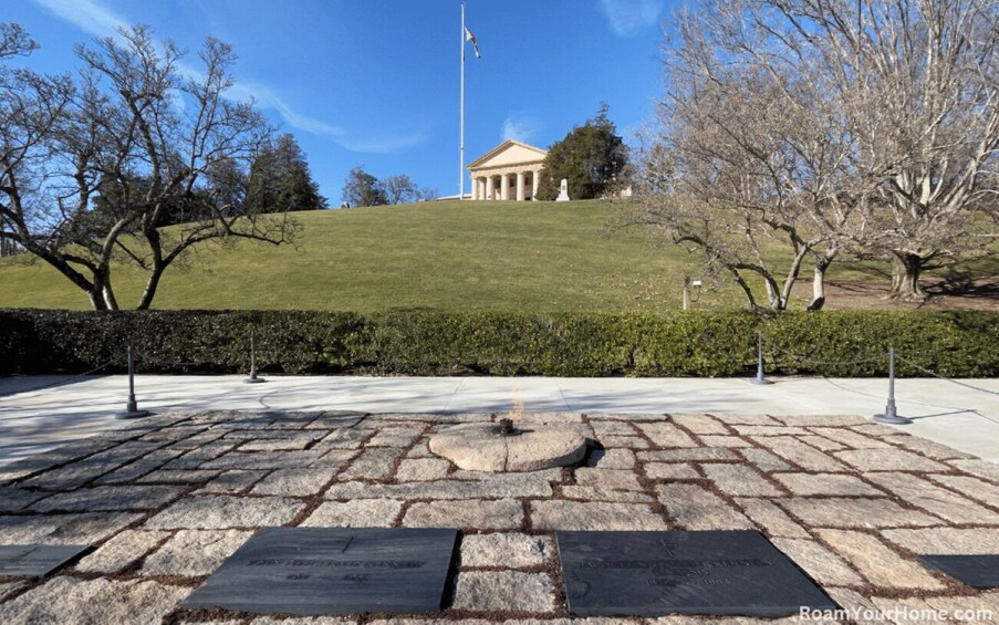 A stone plaza with a flagpole, a building with columns, and a hill with trees. 2