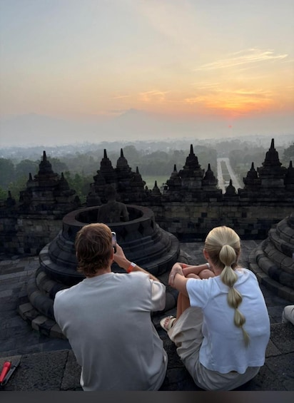 Two people sitting and taking a photo of a temple complex with a sunset in the background. 3