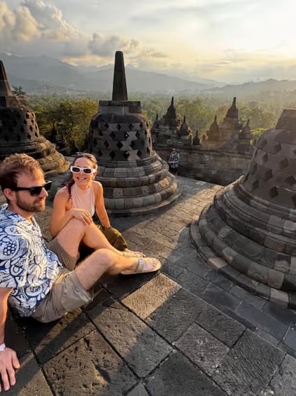 Two people sitting on stone steps overlooking ancient temple structures and a mountainous landscape. 1