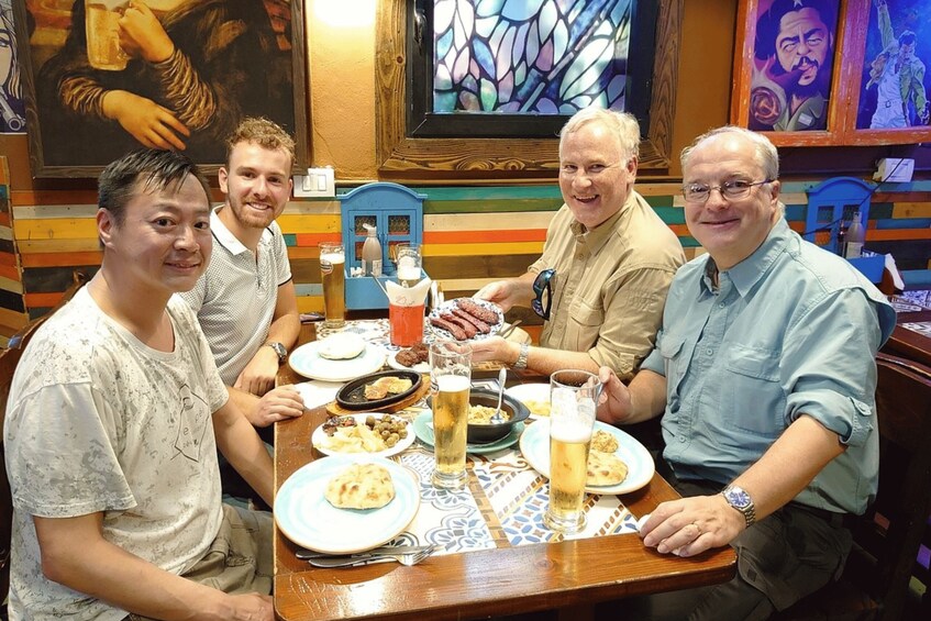 Four people seated at a table with food and drinks, in a room with colorful walls and artwork. 2